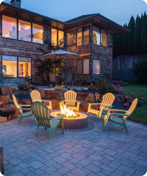Outdoor fire pit with Adirondack chairs on a paved patio in front of a modern home at dusk