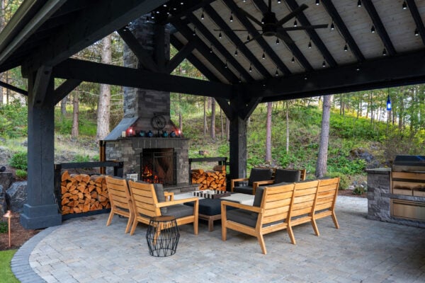 Covered outdoor patio featuring wooden lounge furniture around a lit stone fireplace, with stacked firewood and forest scenery in the background.